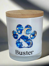 A white jar candle with a wooden lid, featuring a custom stained glass-style paw print design in blue and the name 'Buster' printed on the front.