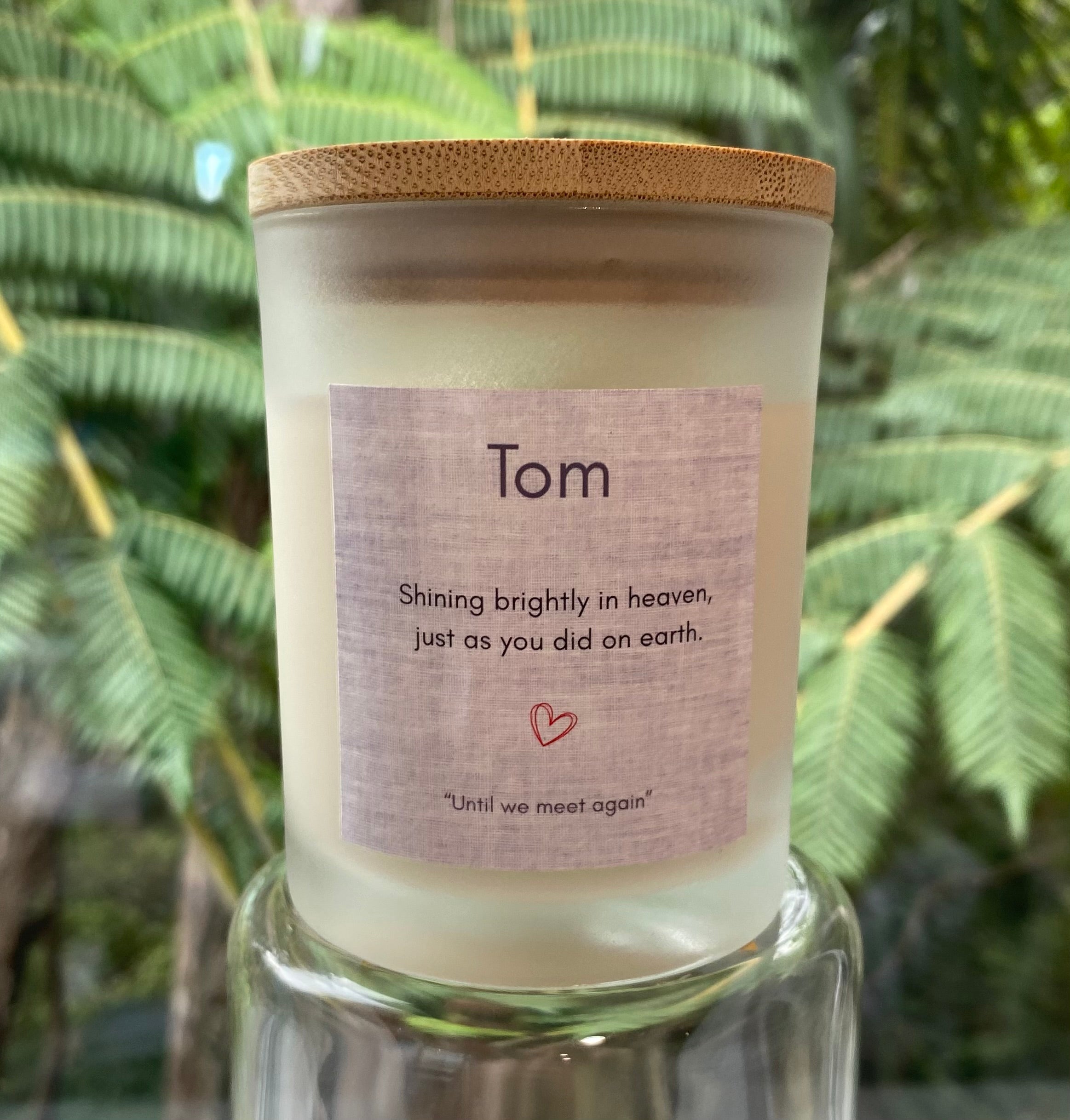 A frosted glass candle jar with a bamboo lid and a custom label reading 'Tom' and 'Shining brightly in heaven, just as you did on earth.' The candle is displayed on a clear glass stand against a blurred background of green tropical leaves.